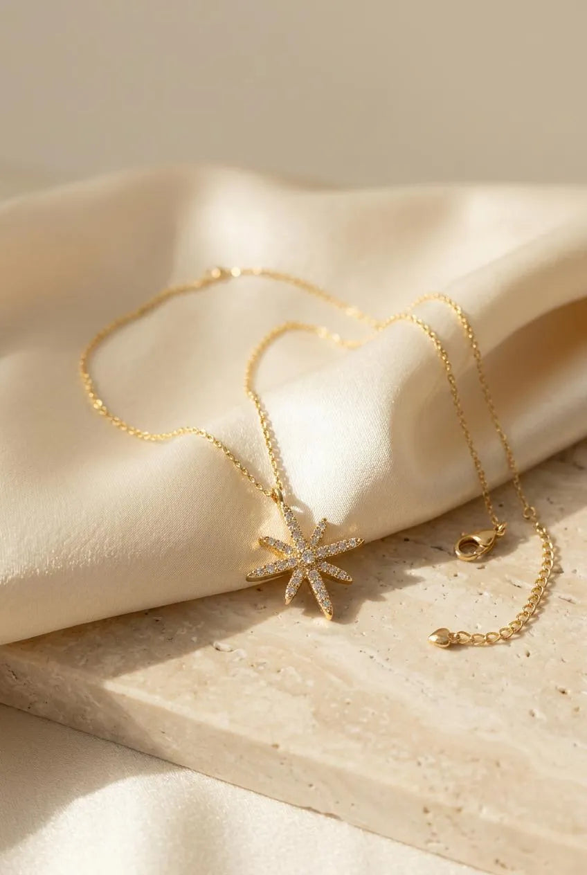 Close up of a luxury gold-plated necklace from Bellora Bloom featuring a celestial radiant starburst pendant paved with sparkling zircon crystals resting on smooth satin fabric.