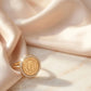 Close up of a luxury gold-plated statement ring from Bellora Bloom featuring a classic engraved Lira coin medallion framed with sparkling zircon crystals resting on smooth satin fabric.