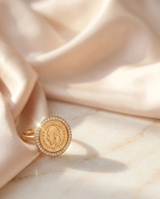 Close up of a luxury gold-plated statement ring from Bellora Bloom featuring a classic engraved Lira coin medallion framed with sparkling zircon crystals resting on smooth satin fabric.