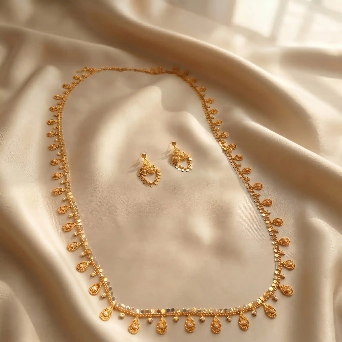 Close up of a luxury gold-plated new arrival long necklace and matching earrings set from Bellora Bloom featuring authentic Khaliji motifs resting on smooth satin fabric and stone display.
