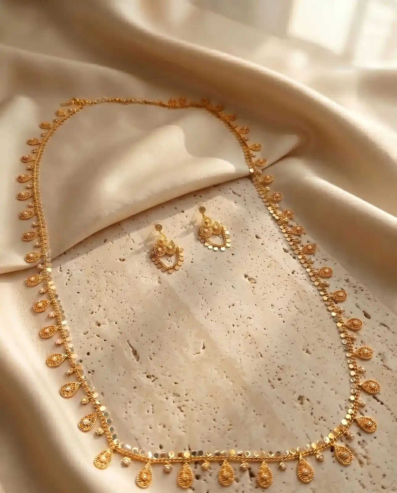 Close up of a luxury gold-plated new arrival long necklace and matching earrings set from Bellora Bloom featuring authentic Khaliji motifs resting on smooth satin fabric and stone display.