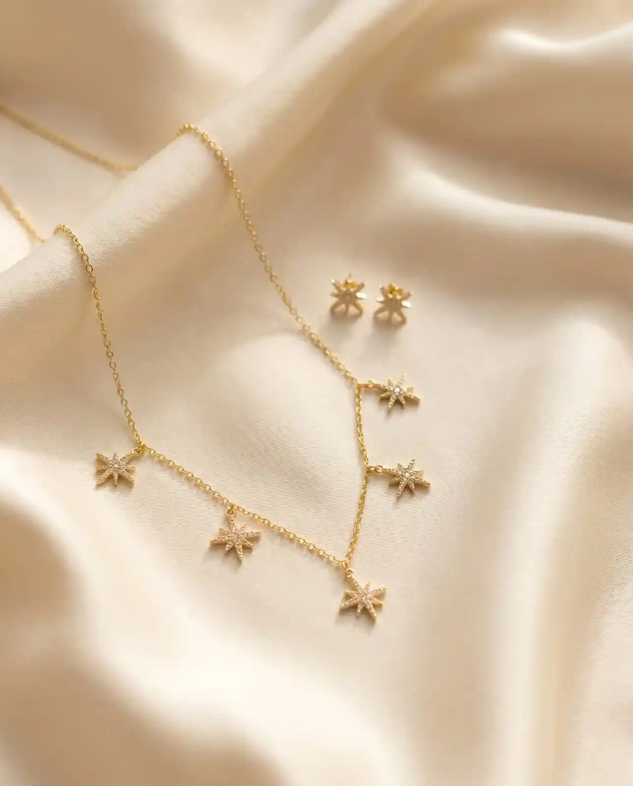 Close up of a luxury 18k gold plated necklace and matching stud earrings set featuring five sparkling star charms paved with clear zirconia on smooth satin.