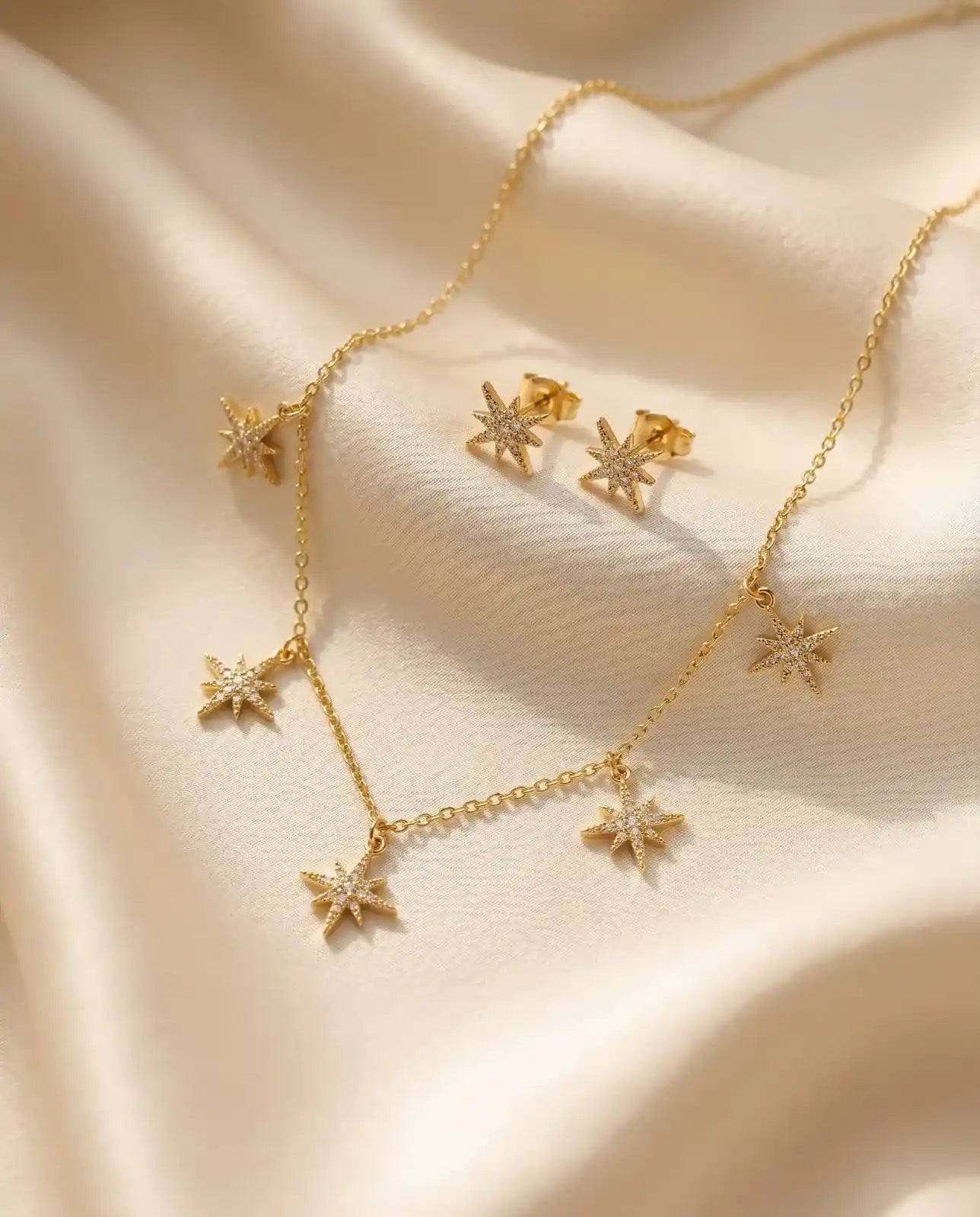 Close up of a luxury 18k gold plated necklace and matching stud earrings set featuring five sparkling star charms paved with clear zirconia on smooth satin.