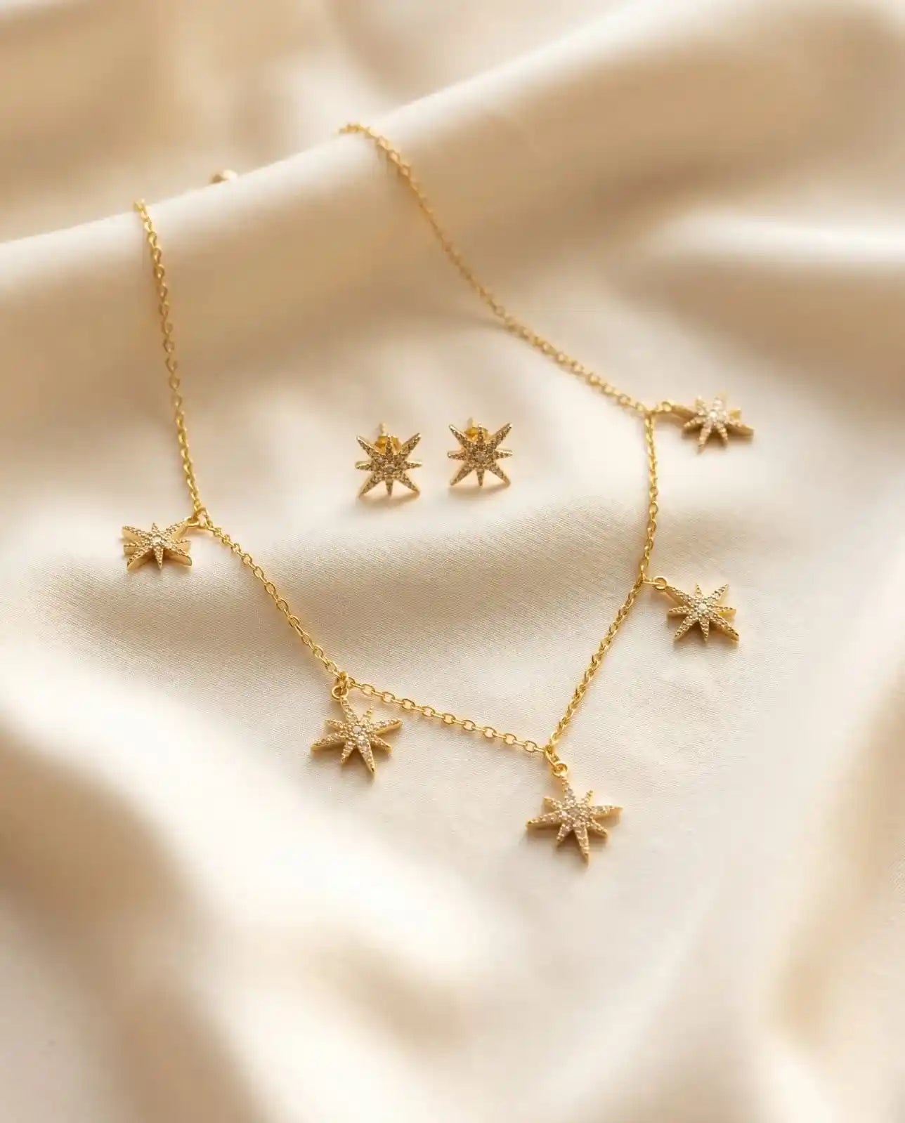 Close up of a luxury 18k gold plated necklace and matching stud earrings set featuring five sparkling star charms paved with clear zirconia on smooth satin.