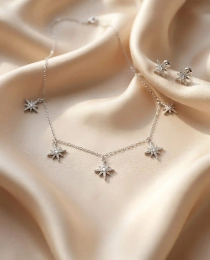 Close up of an elegant silver necklace and matching stud earrings set featuring five sparkling star charms paved with clear zirconia on soft ivory satin.